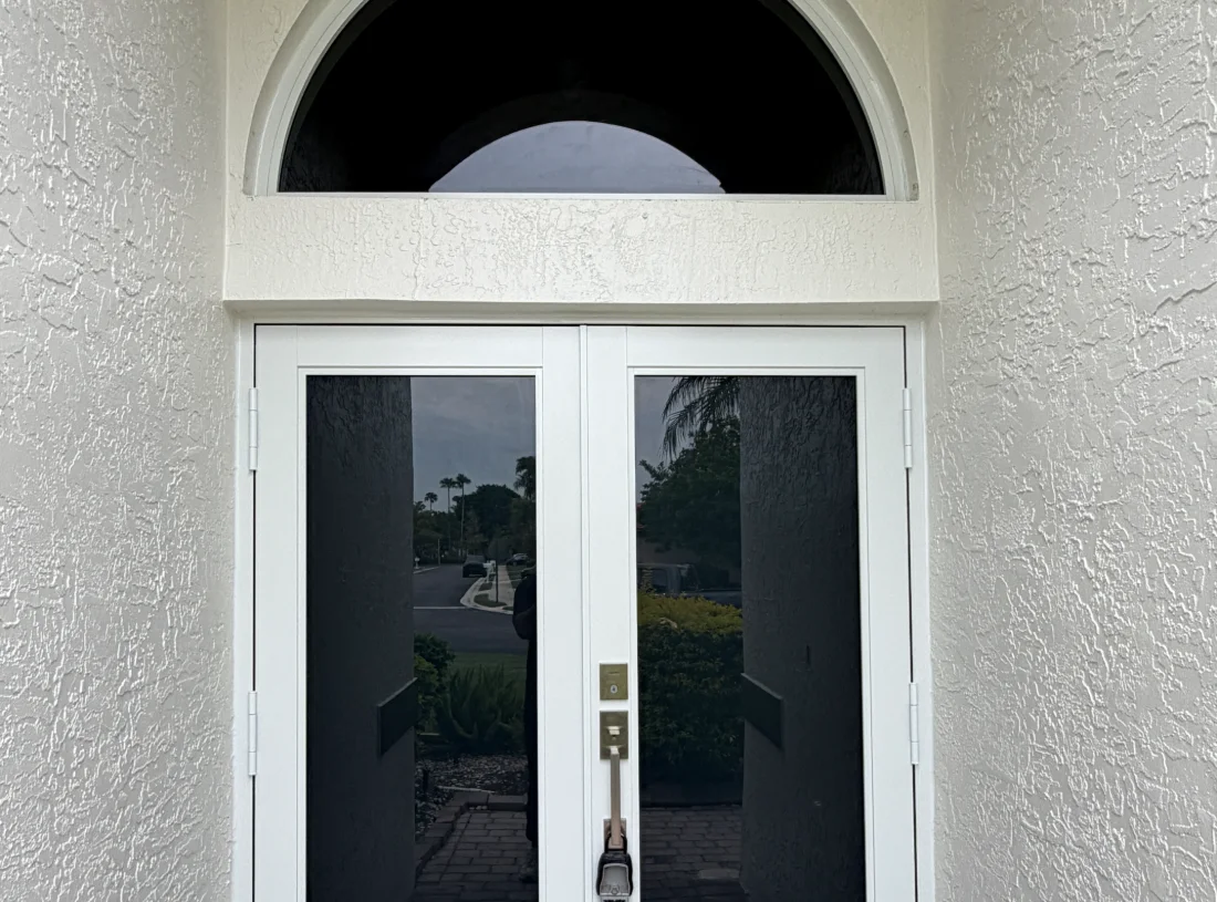 Pair of white-framed impact French doors with full-height glass under an arched transom, seen from the entry path