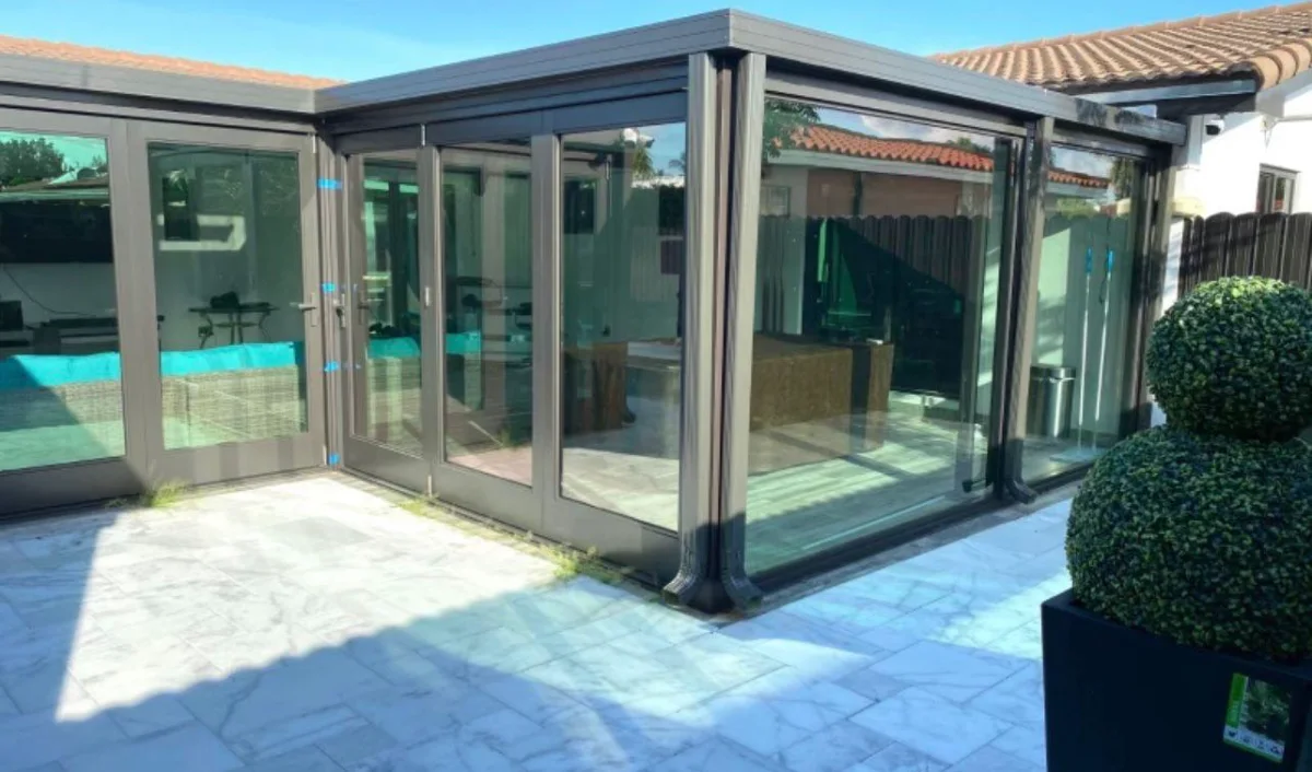 Bronze-framed impact glass patio enclosure with folding doors, opening onto a marble pool deck
